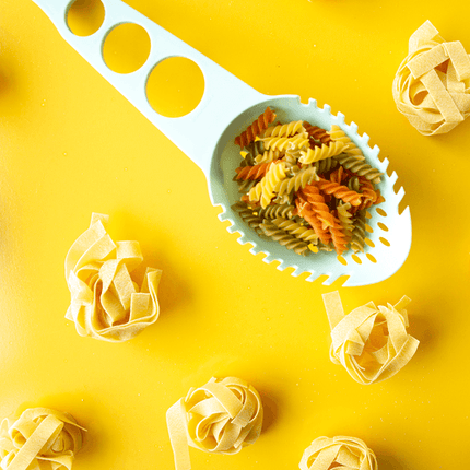 Turquoise spaghettilepel met gekleurde spirelli er in op geel keukenblad met nestjes pasta rondom