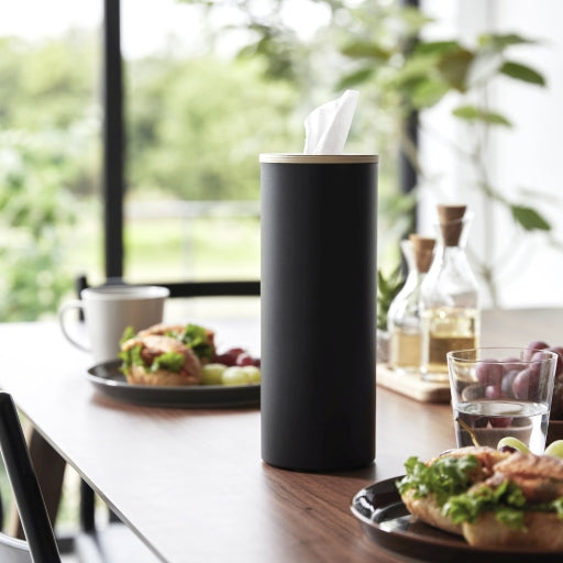 Ronde tissue houder in staal met houten deksel zwart op een houten tafel met een gevuld bord en olijfolie